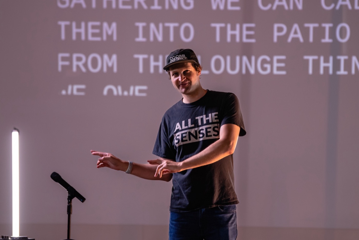 A presenter in a t-shirt reading All The Senses gestures in front of a wall sized projection of live captions in large uppercase type, readable from the back of the room.
