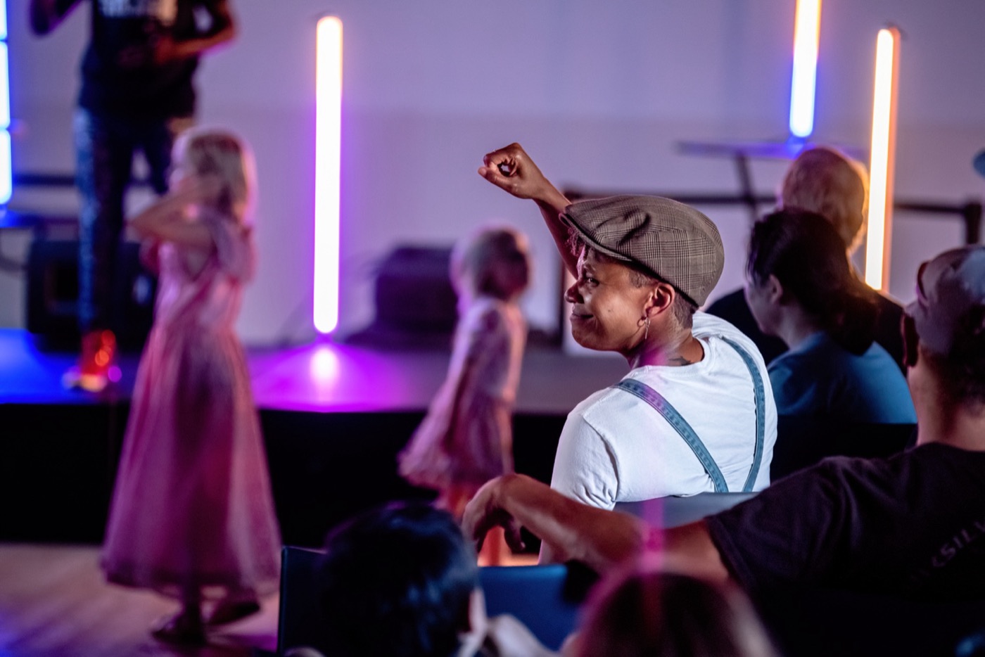 An audience member in a flat cap raises a fist while watching a show, with two dancers on the LED lit stage behind, strongly out of focus.