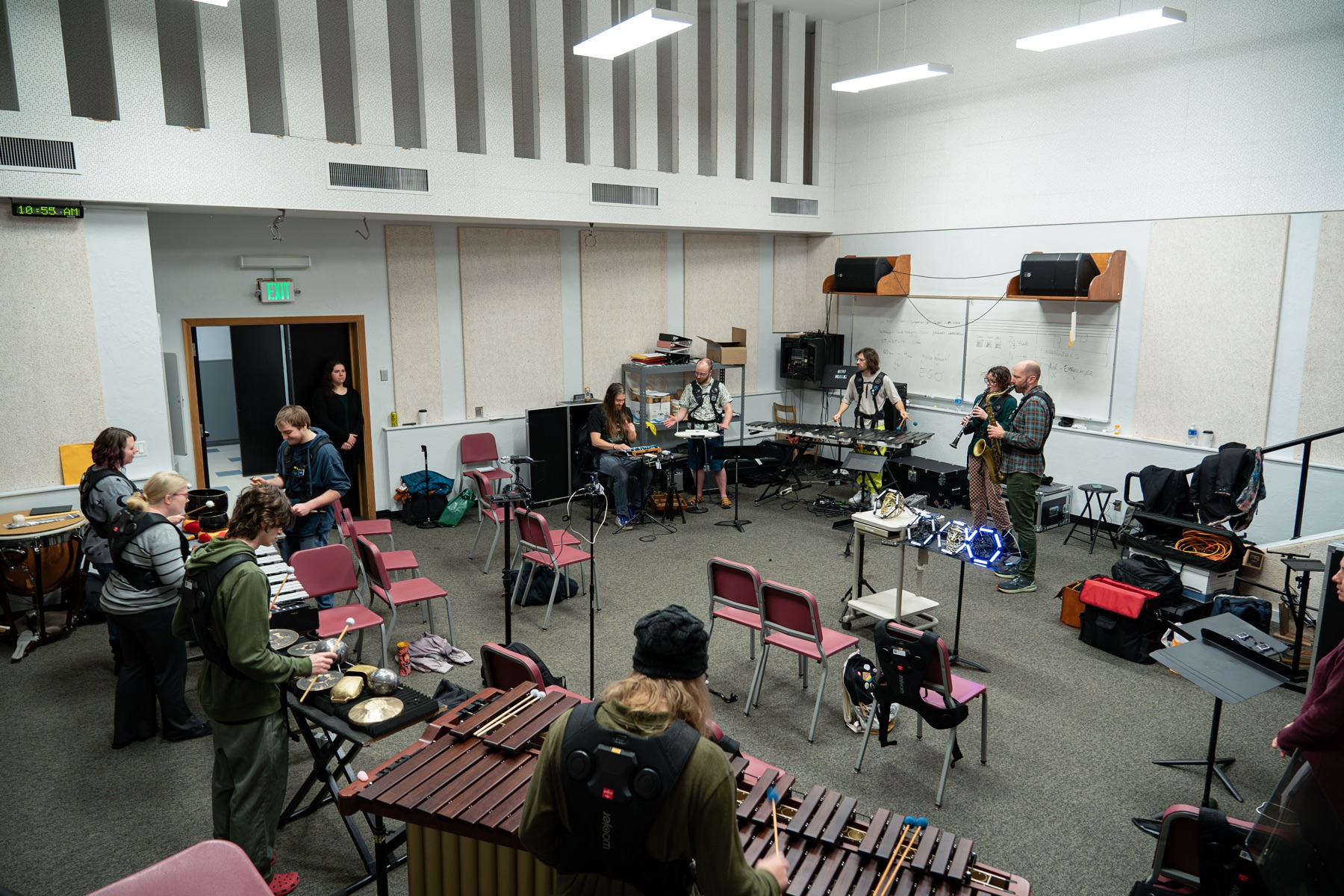 A wide view of a university rehearsal room mid-workshop: about a dozen people wearing haptic vests play marimba, vibraphone, hand percussion, guitar and saxophone, with chairs pushed back and equipment cases stacked along the wall.