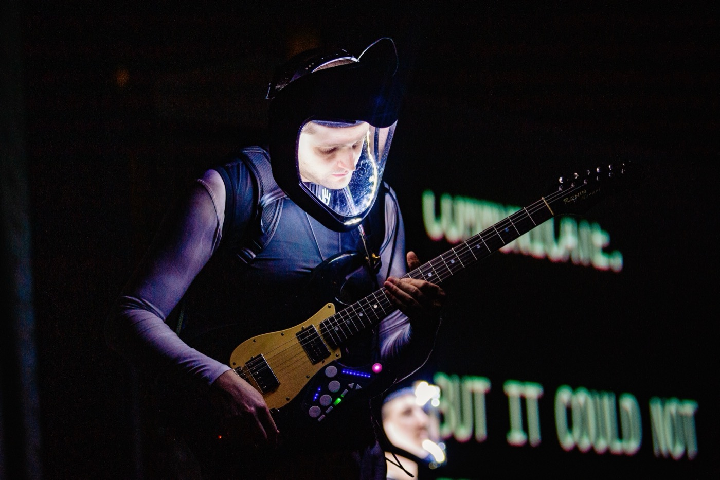 Close view of a performer in a lit-up helmet and a vest, with controls mounted on the instrument and blurred caption text behind.