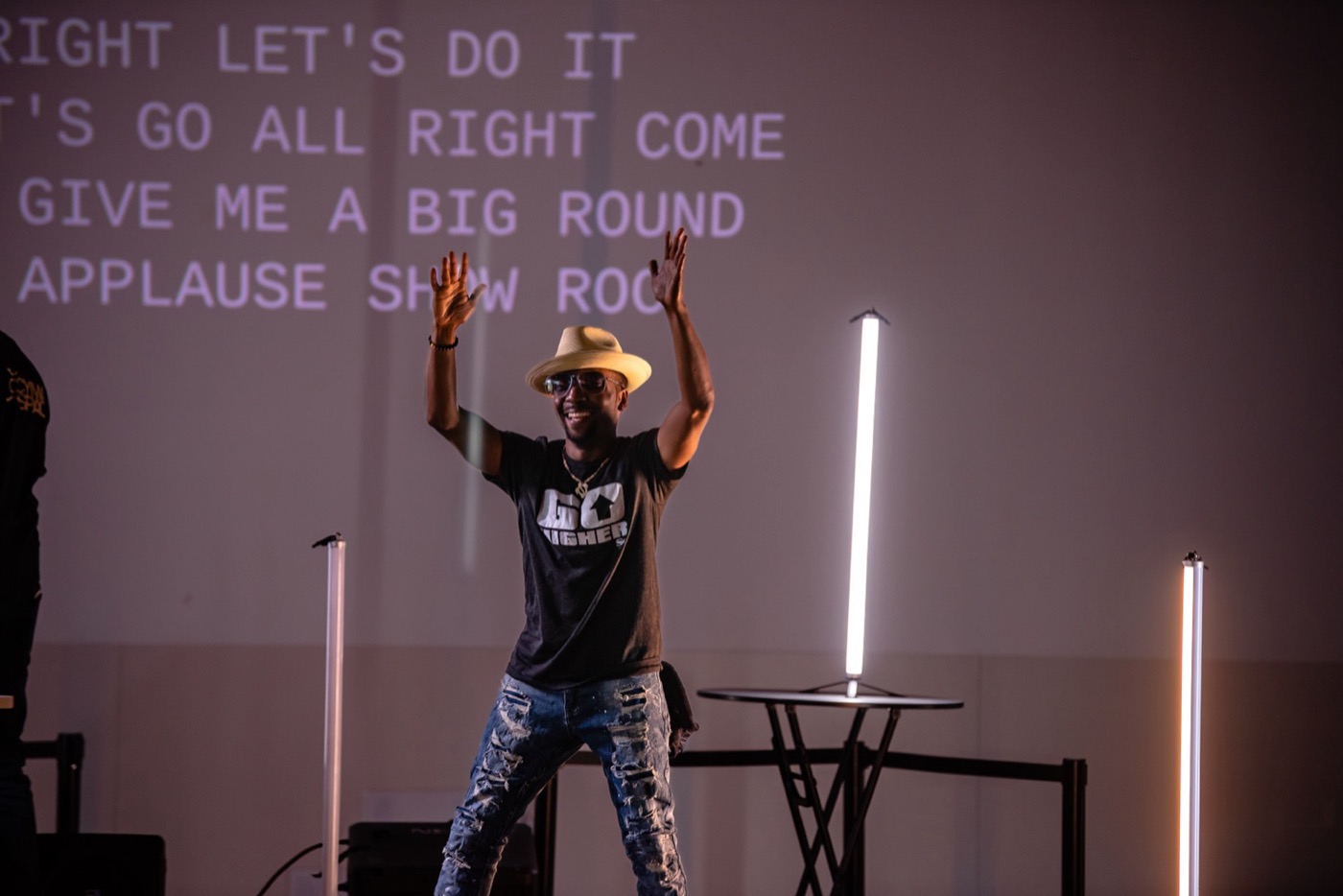 A grinning performer stands with both arms thrown up, and four lines of large capitalized text are projected across the wall behind him at full stage height.