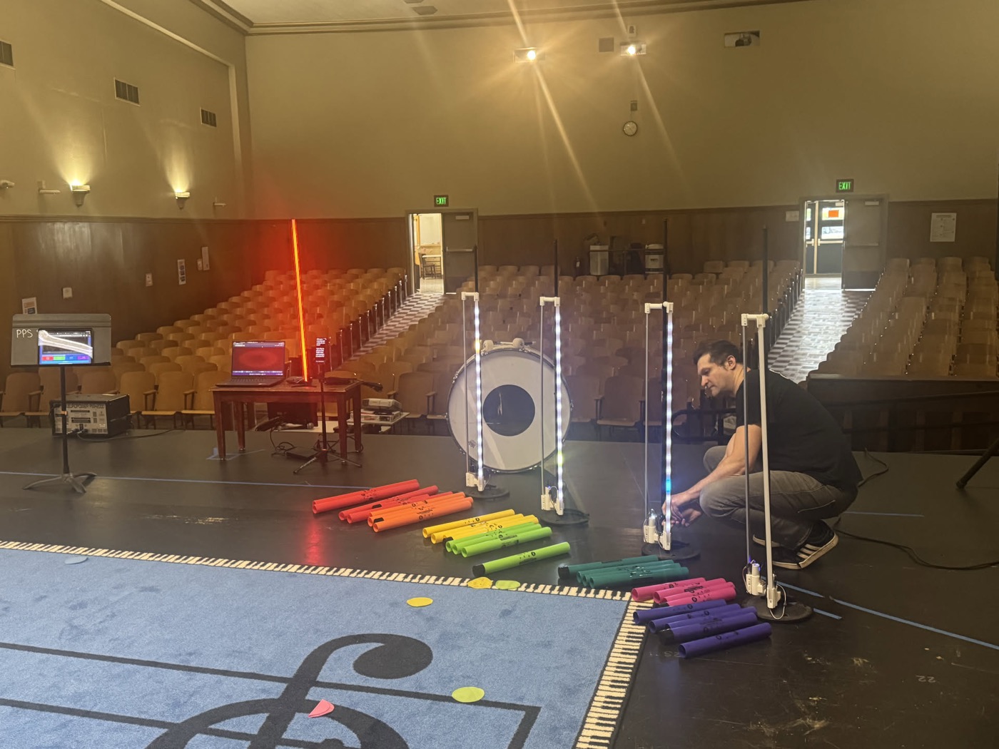 A wide shot of a school stage, instruments lined up along the staff mat and rows of empty seats behind.