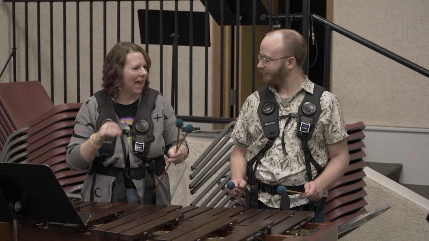 Two participants wearing haptic vests stand at a marimba, each holding a pair of mallets over the bars.
