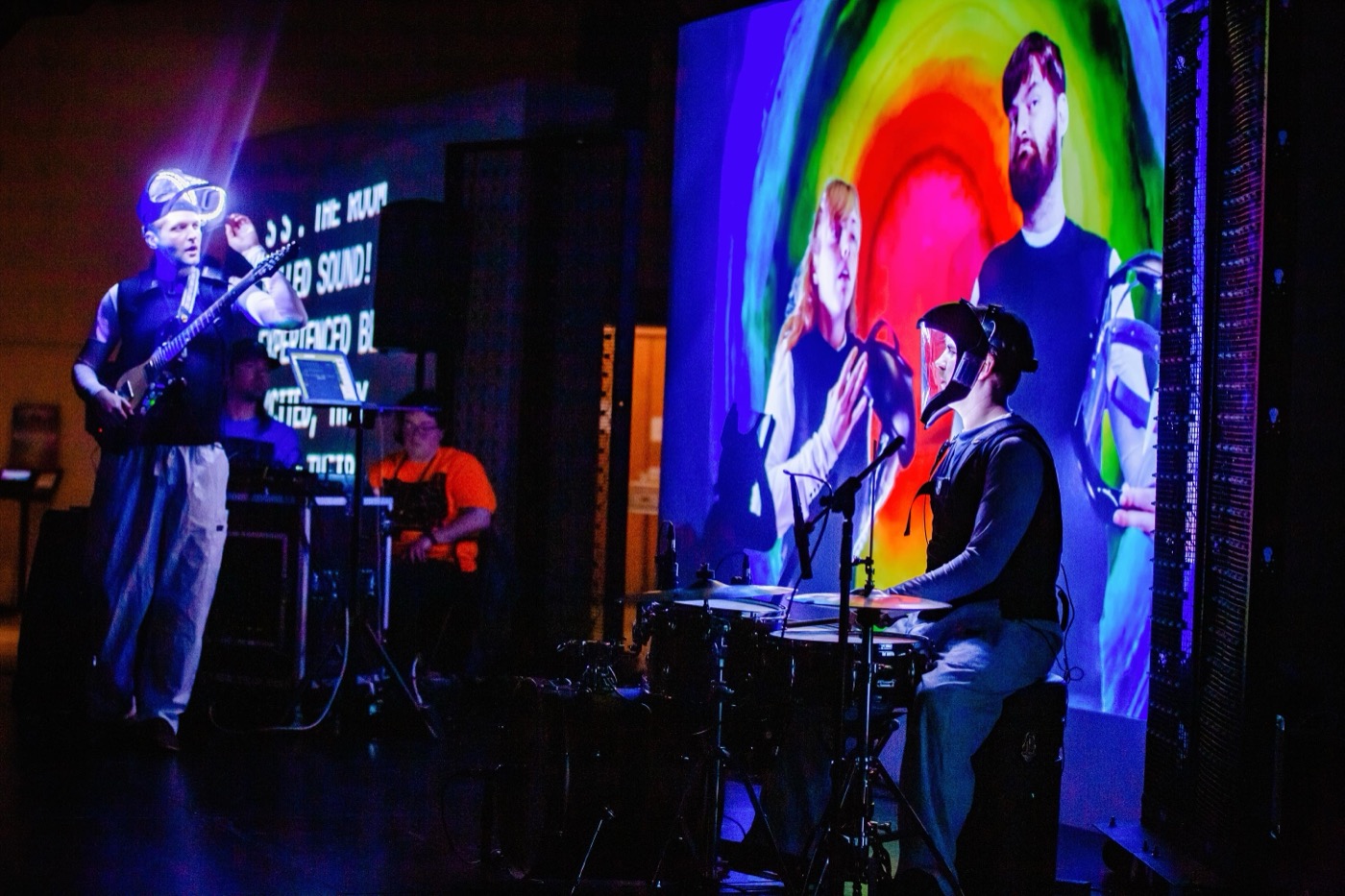 A guitarist and a percussionist play under strong colored stage light, with lines of projected text and bands of rainbow color filling the wall behind them.
