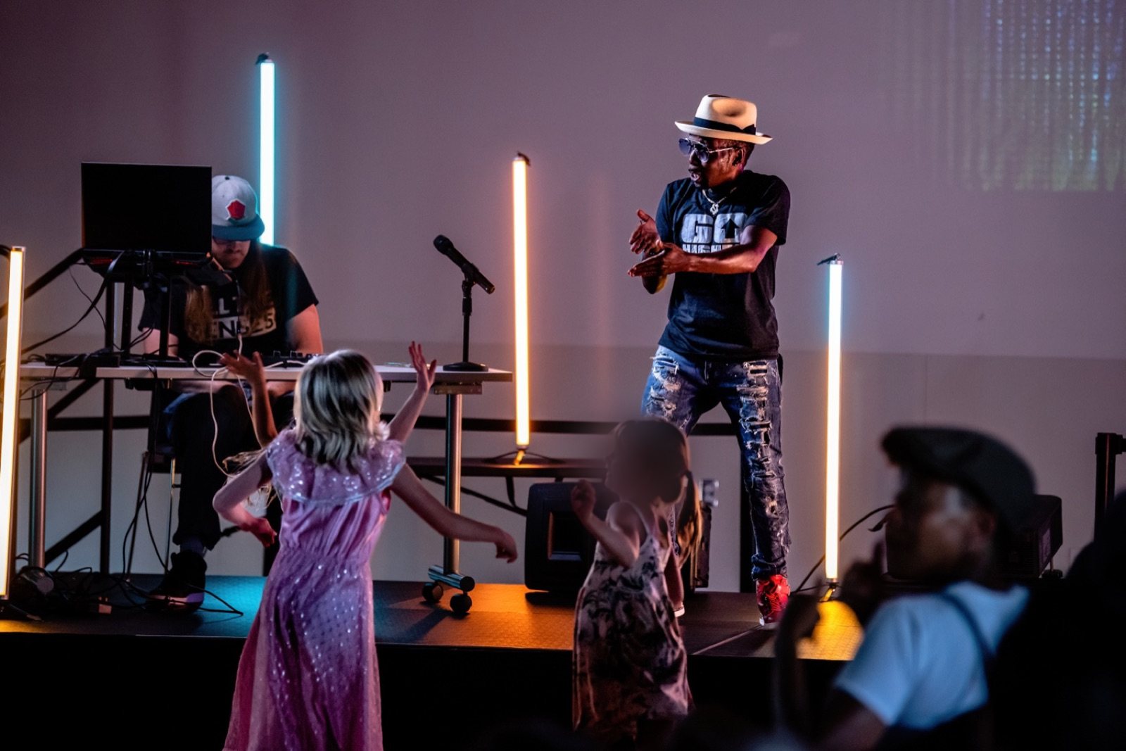 A Deaf performer works a low stage lit by vertical LED tubes while children dance in front of it, one turned away from the camera and one with the face obscured.