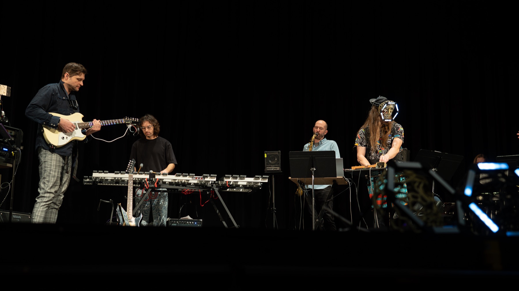 Four musicians perform together on a dark stage with guitar, saxophone, mallet instruments and electronics, wearable lights glowing in the foreground.
