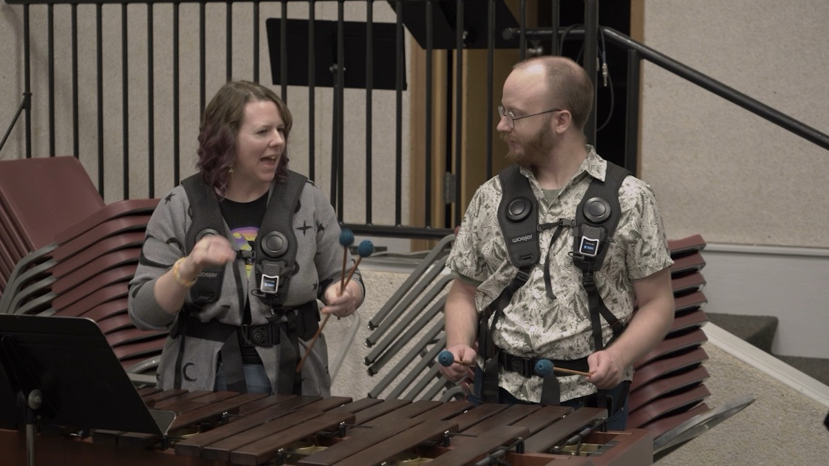 Two people at a marimba, both wearing haptic vests with round drivers across the chest, mallets raised, laughing as they play.